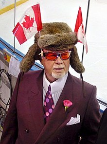Photographie couleur de Don Cherry en costume-cravate dans les tribunes d'une patinoire. Il porte une chapka surmontée de deux drapeaux canadiens.