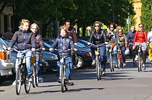Frontale Farbfotografie von einer Gruppe Radfahrern, die in ihrer Alltagskleidung durch eine Straße fahren.