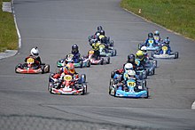 A gaggle of small, low brightly coloured karts underway together on a dark grey tarmac circuit