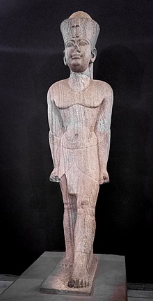 Large light-brown statue of a man striding, wearing the double crown of Ancient Egypt.