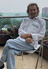 A middle-aged, white man with shoulder-long white hair, a white goatee, white blouse, and light-blue jeans sits cross-legged on a foldable chair on an outdoor balcony. Potted plants are placed in the background and high-rise buildings and trees are visible beyond the balcony.