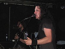 Close-up side view of Joe Duplantier onstage, with Christian Andreu partly hidden in the background