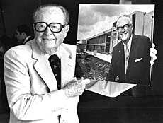 Mitchell Wolfson smiling, posing with and holding a portrait of himself standing behind a Miami-Dade County Community College building.