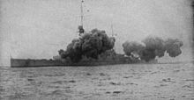 A large warship is partially obscured by smoke from its main guns firing.