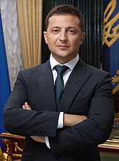 Portrait d'un quadragénaire aux cheveux bruns coupés court, en costume noir, posant bras croisés devant un bureau en bois et l'emblème de l'Ukraine en arrière-plan.
