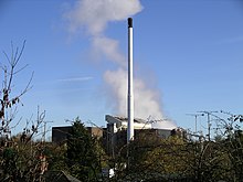 The tops of a row of trees at the foreground obscure a large industrial building. In the centre, reaching to the top, is a white thin chimney and behind this trails white smoke into the sky.