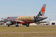 Left side on view of an aeroplane. It is basically silver in colour with the standard logo of the airline. Added to the side of the plane is a large print of 'Powderfinger' in black-bordered yellow to orange-brown colouring. It spans the distance from where the wings join to just before the rear doorway. To the right and just below the band's name, in smaller lettering, is the word 'Sunsets', coloured mostly in brown. Either side of the runway are grassed areas, beyond the jet are buildings – mostly obscured.