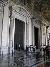 Fotografija prikazuje pogled na predprostor s tremi ogromnimi vrati, ki vodijo v notranjost cerkve. Vrata so uokvirjena s stebri. Tla so iz intardiranega marmorja. Najbližja vrata zapirata dve ogromni starodavni bronasti vratnici. Skupina posluša vodnika, medtem ko ena ženska pregleda vrata.