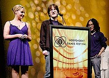 Two men stand behind a podium marked Independent Games Festival, while a woman in a blue dress stands to the left.