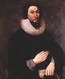 A painting of a man with a stern expression on his face, wearing very dark clothing so that his pale hands show boldly. His hands are placed in front of him, separately, one above the other.