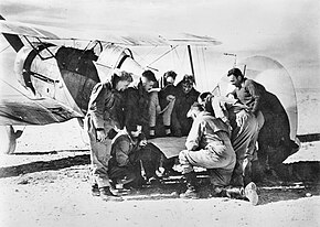 Half a dozen or so men looking at a map on the tail unit of a military biplane