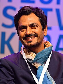 close-up of Nawazuddin Siddiqui wearing a dark purple suit, print scarf, grinning and looking left of camera, with advertisements in background