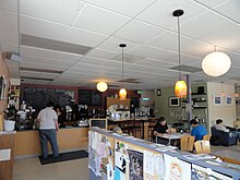 Photograph of the interior of the coffeehouse, with someone at the front cover and patrons seated