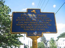 A close-up of a blue and yellow plaque reads: "STEPHEN CRANE: In this park Stephen Crane interviewed men of the famed Civil War Orange Blossoms Regiment and then wrote The Red Badge of Courage, published in 1895."