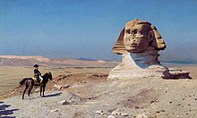 Orang yang menunggang kuda melihat ke arah patung gergasi kepala di padang pasir, dengan langit biru