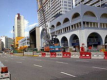 A part of the road has been blocked off with road barriers. Two cranes (one in the forefront) towers over the buildings beside the road.
