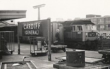 A black-and-white photograph of a railway platform. Two trains stand side by side on the image's right while a sign on the platform reads "Cardiff (General)". A formally dressed gentleman stands on the platform, looking away from the viewer. A briefcase, presumably his, rests on a small trolley beside him