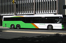 Xe buýt màu xanh nhạt, cam và trắng dừng trước tòa nhà nhiều tầng.