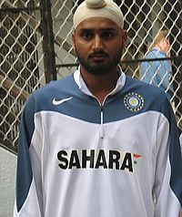 A Black-bearded man wearing a white turban stares directly at the camera. He is wearing a grey-blue and white shirt with the colours of the Flag of India and the word "SAHARA" on the shirt's chest area.