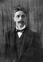 Head and torso of standing middle-aged white man, in a dark suit, with full head of wavy dark hair, looking to the side. He has a neat, medium-sized moustache