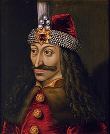 Painting of a richly dressed Caucasian man's head and shoulders. He has long, curly black hair and a moustache. He is wearing a red hat with a wide band of pearls and a gold star and red gemstone on the front. He is wearing a brown fur cape, over a red and gold top.