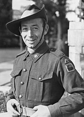Black-and-white photo of a smiling, clean-shaven man wearing combat fatigues and a slouch hat. He is standing hands on hips with his hands gripping his light-coloured belt.