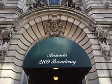 The entrance to the Ansonia. A green awning with the words "The Ansonia, 2109 Broadway" is placed atop the door. The doorway is surrounded by limestone blocks, and there is a limestone face carved above the doorway.