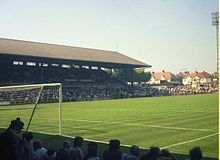 A colour photograph, taken from behind a goal net, of a roofed English-style grandstand, filled with people