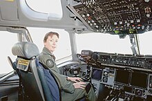 Colour photo of a woman wearing a green flight suit sitting inside an aircraft cockpit