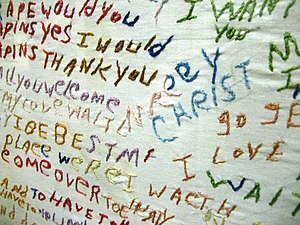 A white cloth with seemingly random, unconnected text sewn into it using multiple colors of thread