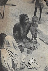 Old photograph of a woman squatting and tiny, emaciated toddler standing on a sidewalk. The woman is shirtless but squatting to conceal her breasts. The toddler is wearing rags.
