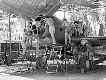 Black and white photo of a single-engined military monoplane being serviced by eight men wearing only shorts and shoes. The aircraft is on the ground below netting.