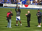 Four men standing on a grass football pitch. The man second from left, wearing a grey top, white shorts and white socks, is holding a trophy above his head. Spectators wearing blue or black tops are visible in the background.