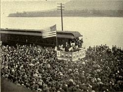 A dramatic political scene. Beside a river stands a podium, on which a flagpole flies a huge American flag. Beneath the flag stands a candidate in a dark suit addressing an impressive crowd which takes up most of the photograph. Not only the quayside but a ferry beside it on the water are packed full of people listening intently.