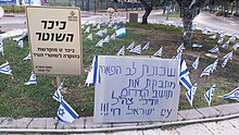 Signs in Hebrew in front of dozens of small Israel flags planted in the grass.