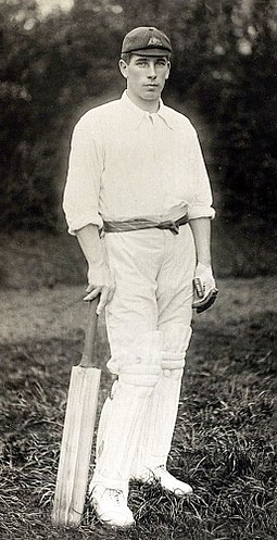 A man standing in cricket whites holding a cricket bat by his side