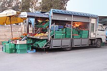 La photographie couleur concerne une camionnette débâchée. L'arrière est déplié en comptoir pour présenter les fruits et légumes à la vente dans des cagettes plastiques vertes et des cageots en bois. Des oranges, des pommes jaunes, des pommes de terre, des tomates, des salades, des oignons... constituent la marchandise.