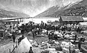 Första guldfyndet görs i Klondike denna dag för 128 år sedan, och utlöser den efterföljande guldruschen. Fotografiet visar ett guldgrävarläger i Dyea i Alaska under guldruschens dagar.