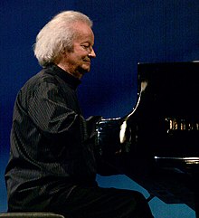 Photographie en couleurs d'un pianiste âgé, habillé de noir, à son instrument.