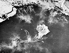 Black and white photograph of a warship moored near a snowy shore viewed from the air. Smoke is issuing from the warship.