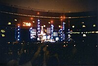 An elaborate concert stage set bearing a logo that reads "Zoo TV", set in a dark stadium. Towers reach into the nighttime sky, illuminated in blue with red warning lights on top.