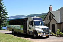 A small bus painted with a mural is parked in front of a house near a lake. Lettering on the bus door says, "Portland Water Bureau: From Forest to Faucet".