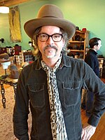 A man with a goatee stands in a room with his left hand on his hip. He is wearing a brown fedora, dark-brown glasses, a pocketed dark-teal shirt, and a leopard-print scarf.