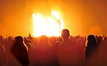 Fotografie nocturnă a unui foc arzând înconjurat de siluete de oameni.