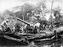 Photographie d'un imposant canon sur un affût rudimentaire tiré sur un pont sommaire en travers d'un petit cours d'eau. Des hommes en uniformes clairs de style coloniaux se tiennent à proximité.