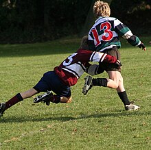 A child running away from camera in green and black hooped rugby jersey is being tackled around the hips and legs by another child in opposition kit.
