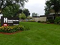 The Westonka Library, a modern off-white and black building located among foliage. Its welcome sign is in front and off to the left, in a bed of yellow flowers.