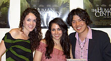 The heads and upper bodies of three people side by side, posing for the camera smiling. At the left, and centre are two white women in their mid-twenties, both with long dark-brown hair. The woman on the left is wearing black and green clothing, and the woman in the centre is wearing pink. At the right is a Japanese man in his early thirties, with dark centre-parted hair. He is wearing a pink open-collar shirt, and a black suit-jacket.