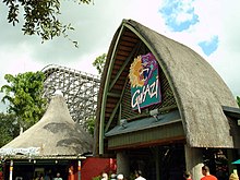 The original Gwazi entrance is viewed in 2006. The thatched roof containing the original Gwazi logo depicting the clash of the yellow-orange tiger and blue-purple lion is seen above the word "Gwazi". The entrance is located on the right, with another thatched roof structure on the left. The Lion's lift hill is seen above the left thatched roof with tree foliage in the foreground.