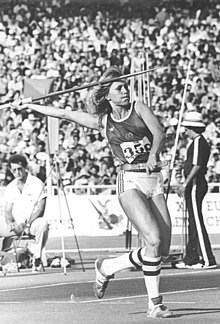 A light-skinned woman, with her arm extended backwards, holding a javelin